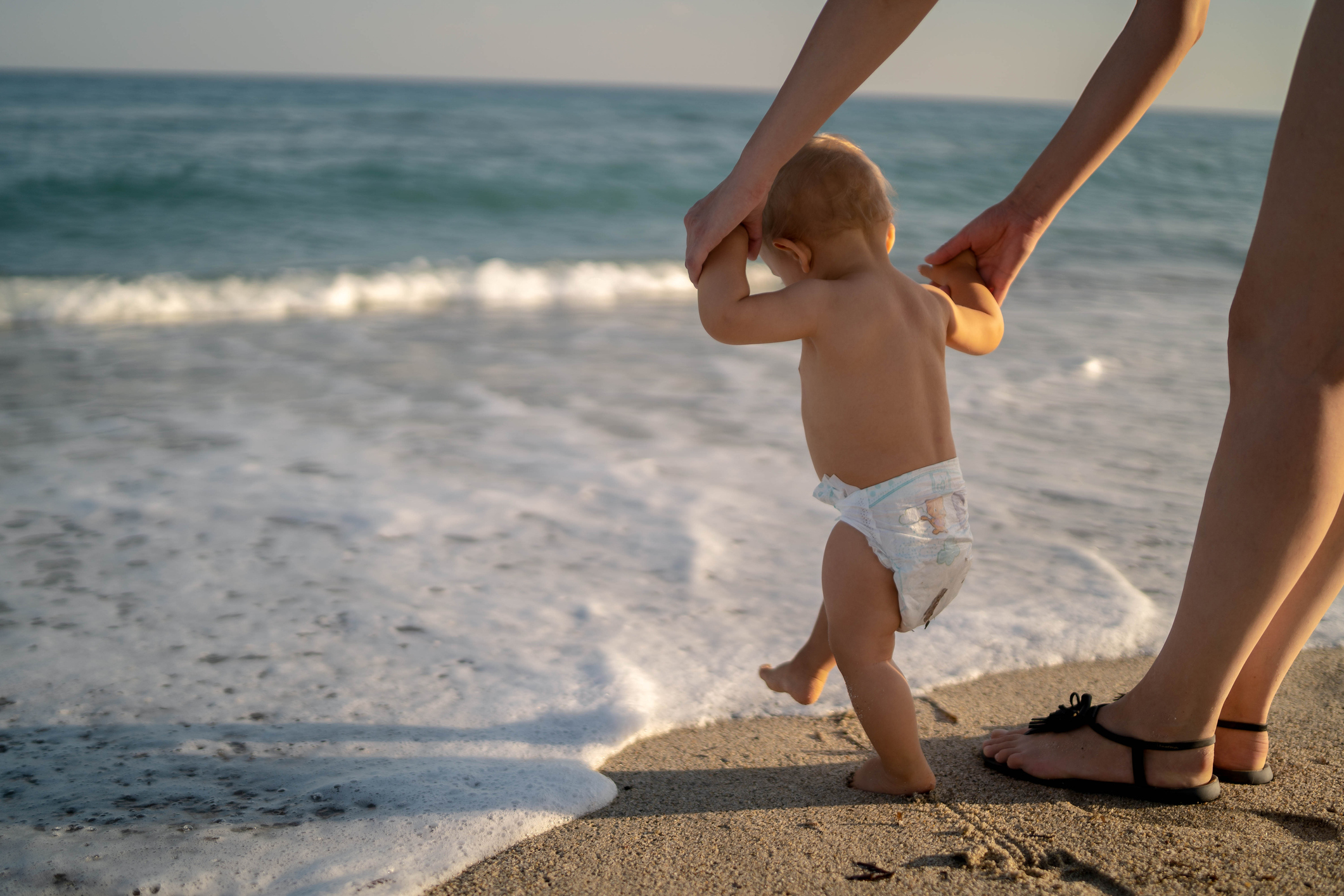 An adult helping a baby in a diaper take steps on the beach near the ocean