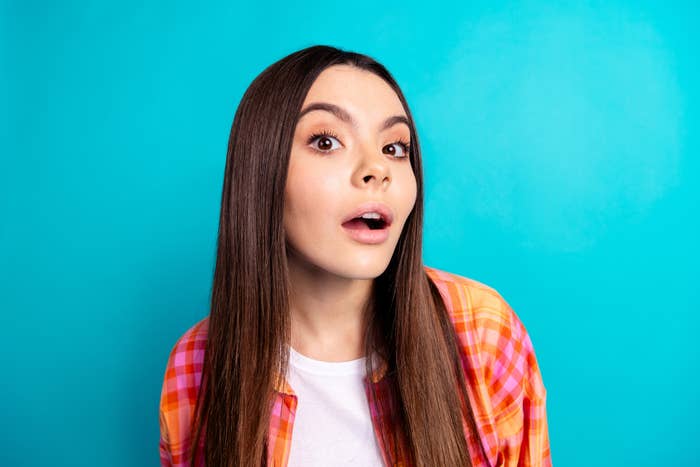 A woman with long, straight hair wears a shirt and a plaid jacket, posing against a plain background with a surprised expression