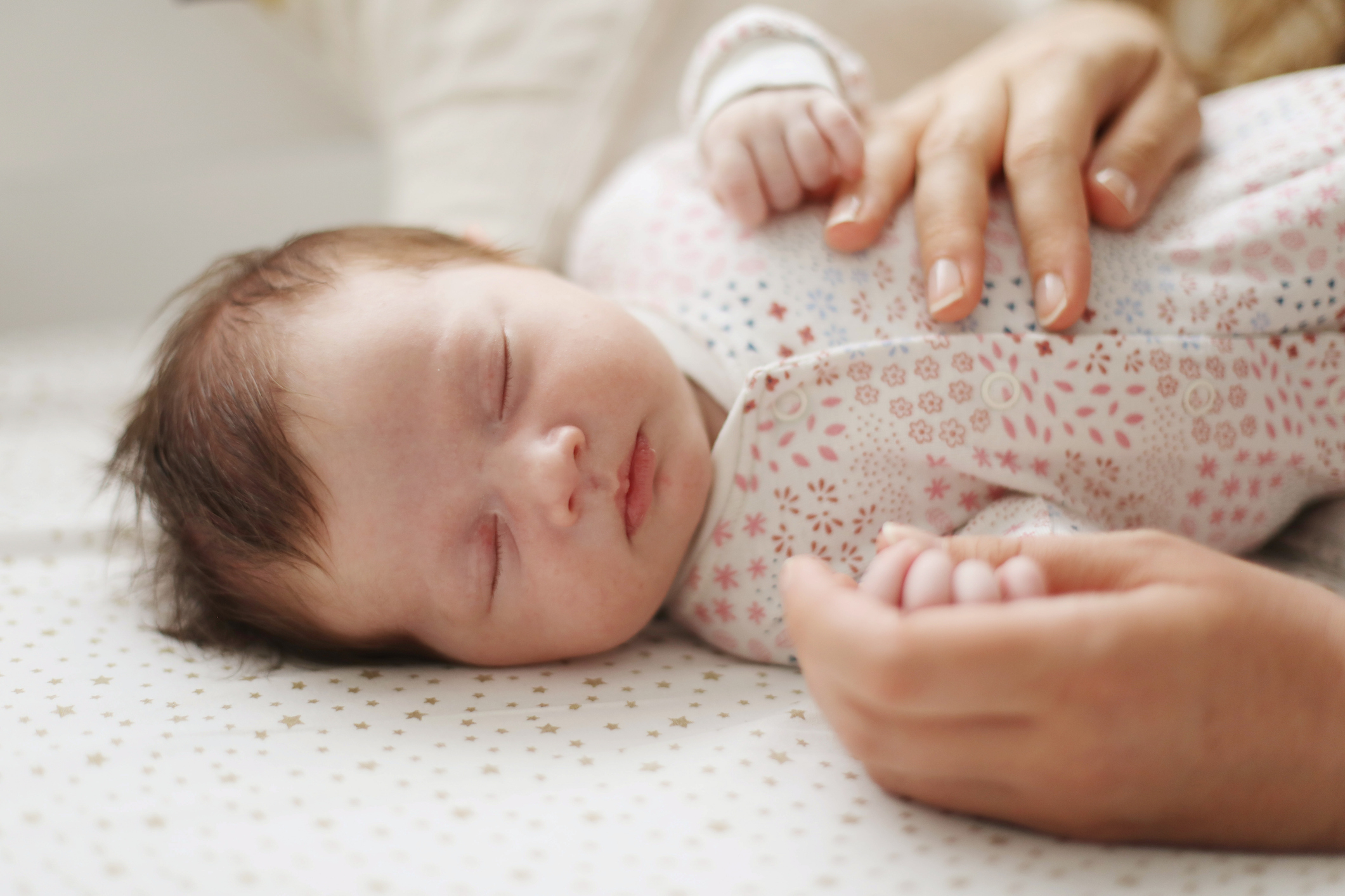 A sleeping baby dressed in a patterned onesie, with an adult's hand gently holding the baby's hand