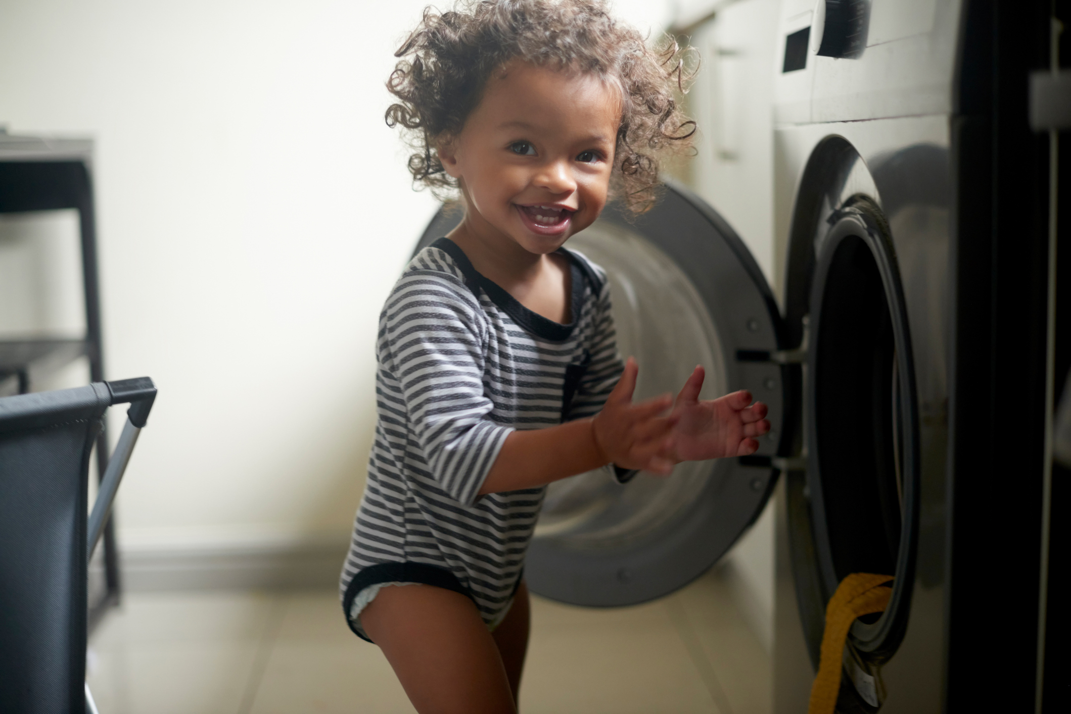 A smiling young child with curly hair in a striped onesie reaches towards an open washing machine