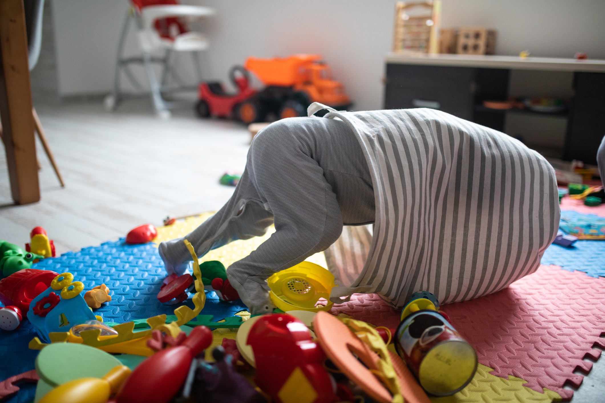A child playing in a room filled with toys, head and shoulders inside a striped cloth bin, surrounded by colorful toy cars, blocks, and play mats