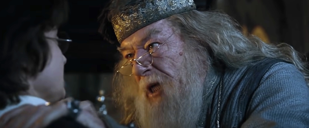 Albus Dumbledore, wearing a detailed hat and glasses, firmly grips Harry Potter by the shoulders during an intense scene in a Harry Potter movie