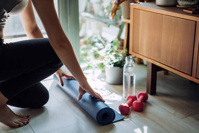 Person kneeling on the floor while rolling up a yoga mat, next to pink dumbbells and a water bottle, inside a cozy, bright room with plants and modern furniture