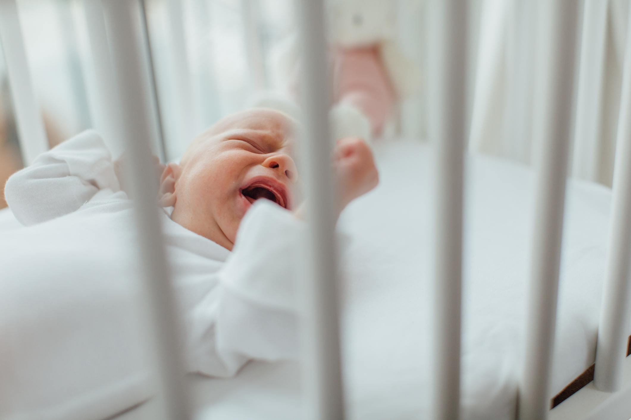Newborn baby crying in a white crib with a stuffed animal in the background
