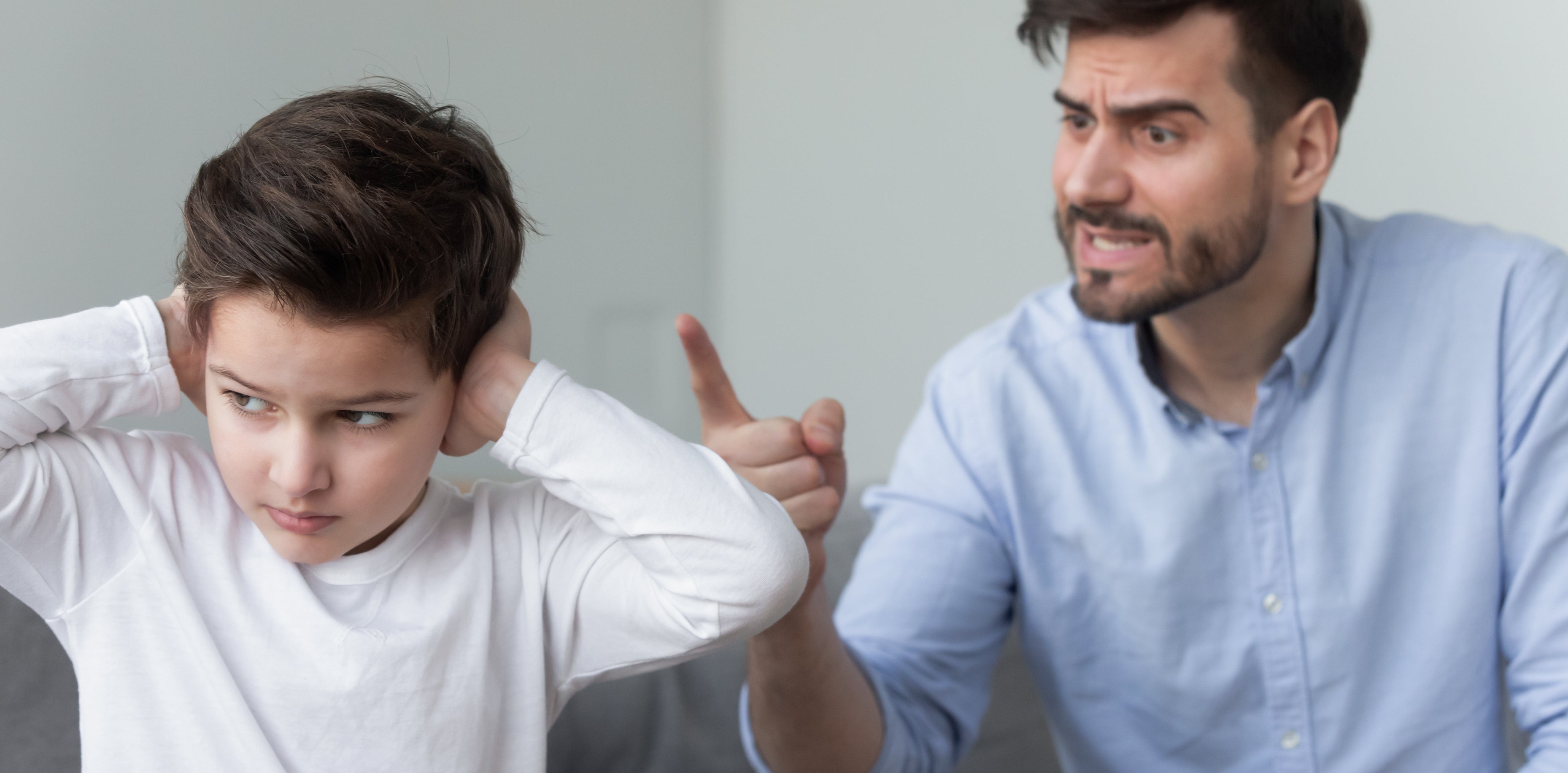 A man in a light blue shirt is pointing and seemingly scolding a boy who has his hands over his ears