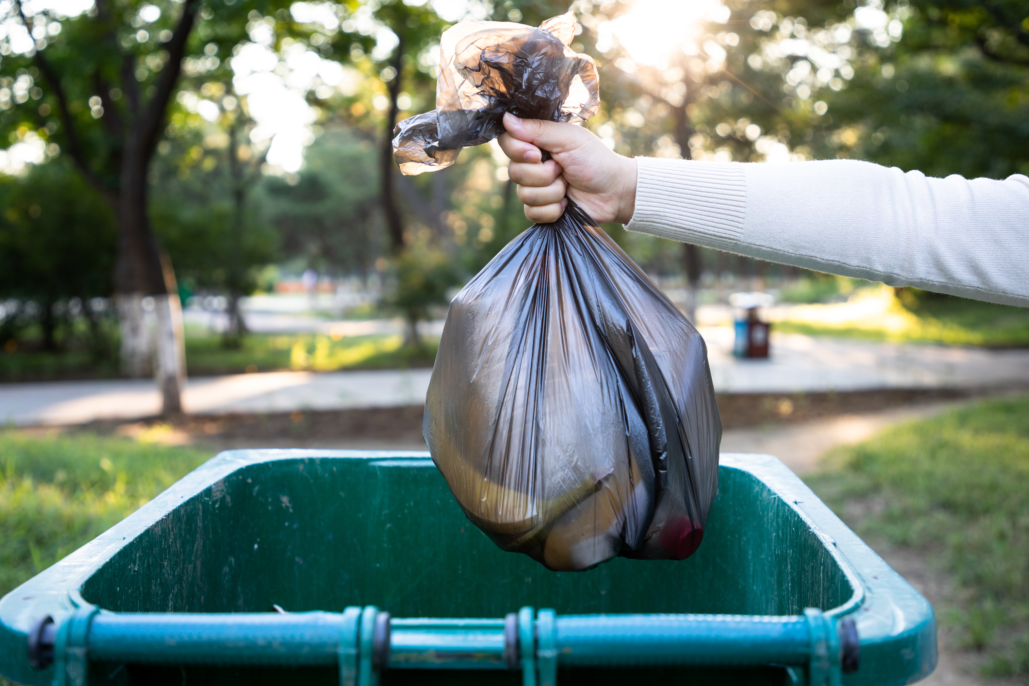 A person holding a tied black plastic garbage bag over a green outdoor trash bin with a park in the background