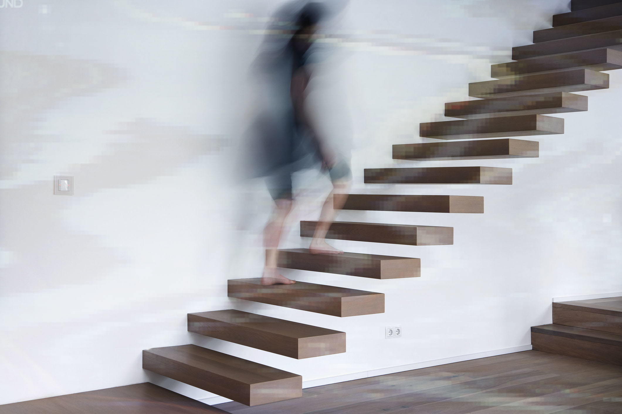 Blurry image of a person walking upstairs on a modern, floating staircase attached to a plain wall. The person's identity and details are indiscernible