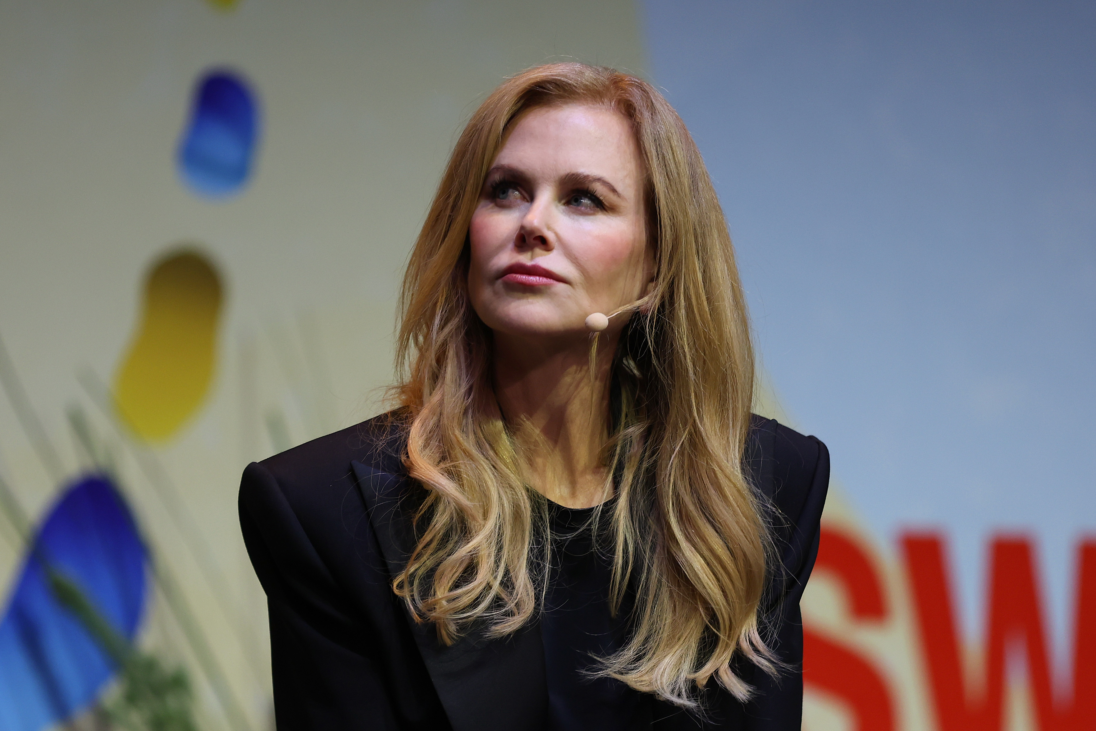 Nicole Kidman is seen at a speaking event, wearing a blazer. She is looking slightly upwards, with a microphone attached to her ear