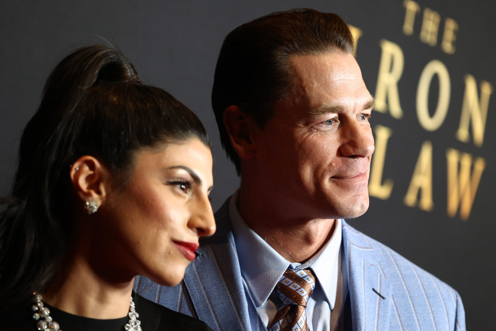 John Cena and Shay Shariatzadeh pose together at an event; John wears a light-colored suit and patterned tie, Shay wears an elegant black dress with jewelry