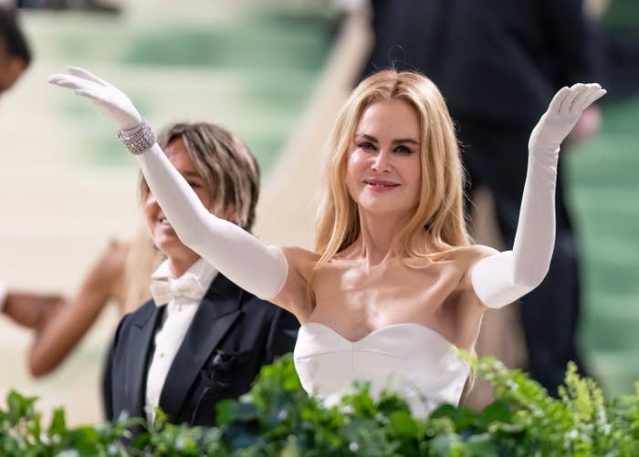 Nicole Kidman is smiling and waving at an event, wearing a strapless gown and long gloves. A person in a tuxedo is partially visible next to her