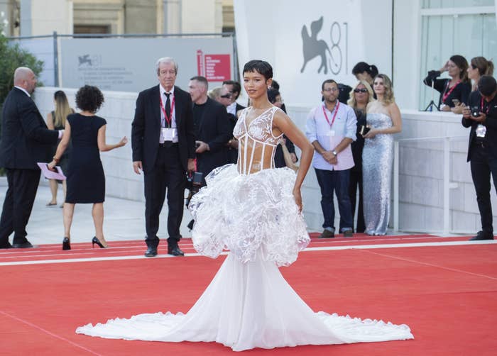 Taylor Russell is wearing an intricate white gown with a flowing train on the red carpet at the Venice Film Festival, surrounded by onlookers and photographers