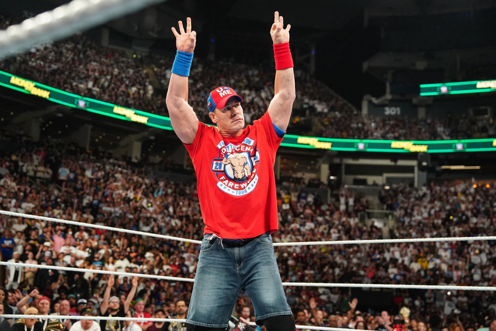 John Cena stands in a wrestling ring during a WWE event, wearing a T-shirt, denim shorts, and a hat, raising three fingers with both hands