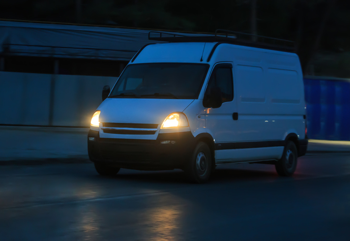 A white delivery van driving down a road at night with its headlights on
