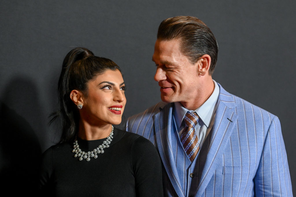 John Cena in a striped suit and Shay Shariatzadeh in a dark dress with a jeweled necklace, smiling at each other on the red carpet