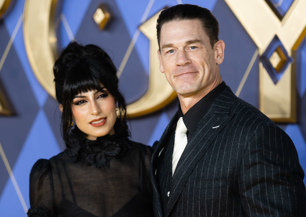 Shay Shariatzadeh and John Cena on the red carpet. Shay wearing a high-neck black dress with sheer sleeves, and John in a pinstripe suit with a white tie