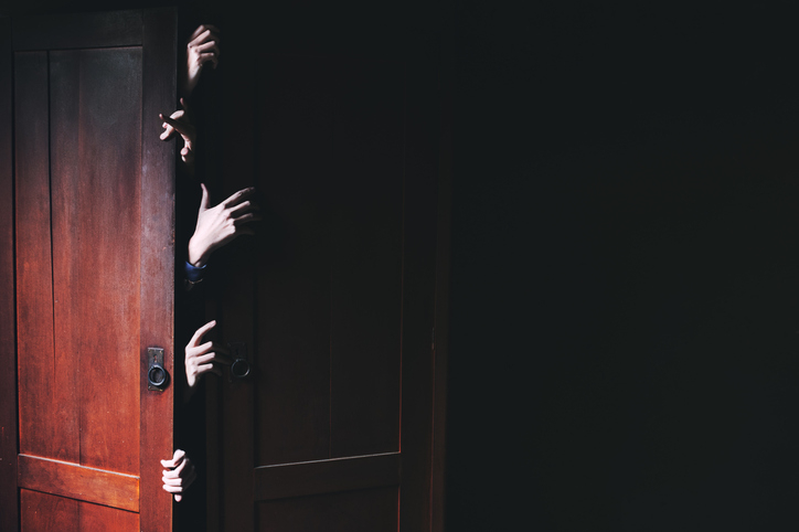 Multiple hands reaching out from a slightly open wooden door, emerging dramatically into a dark space