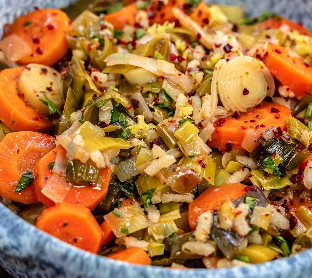 A bowl filled with a vegetable medley dish, featuring cooked carrots, leeks, and herbs, garnished with red pepper flakes. Lemons are in the background