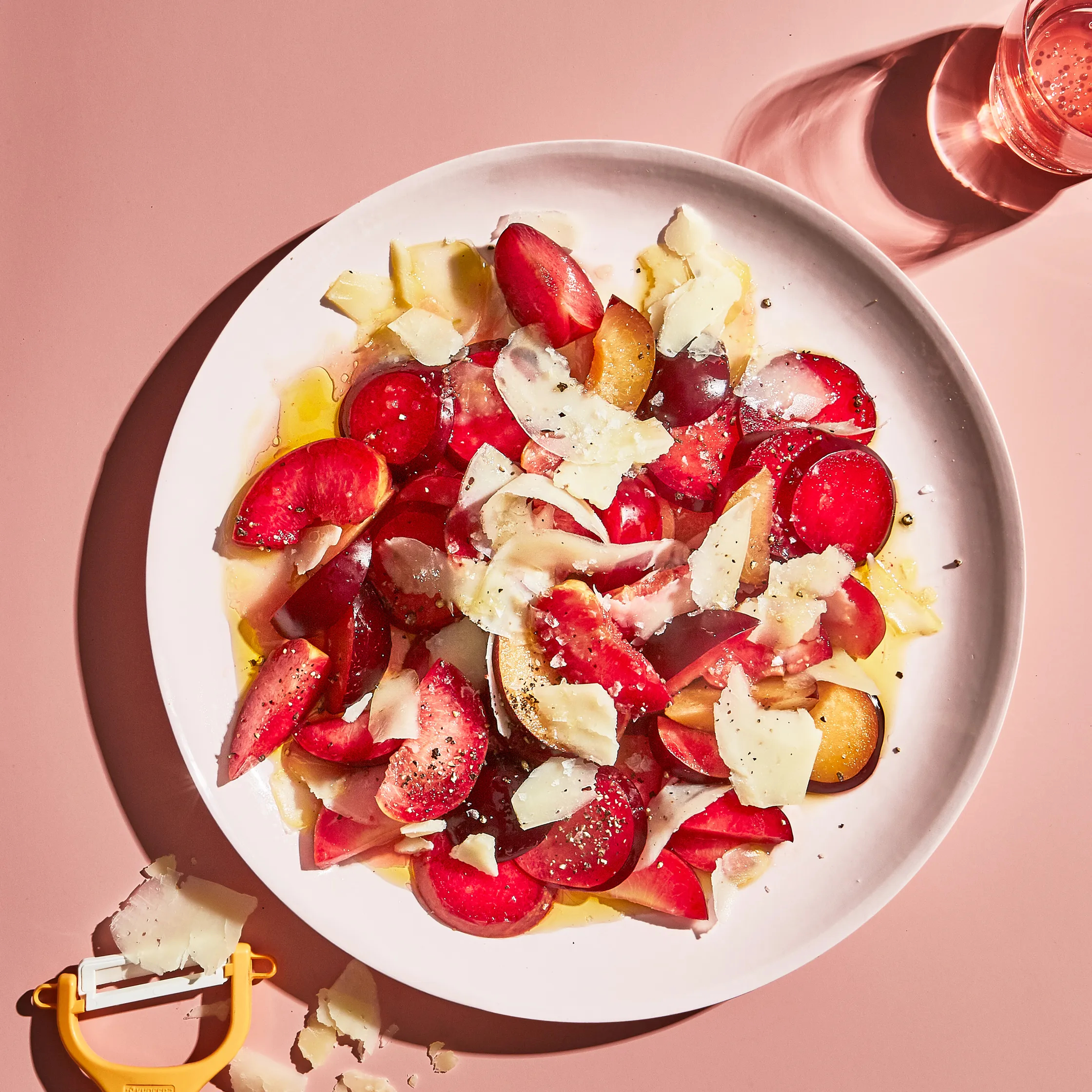 A white plate with a neatly arranged salad of sliced plums, thin cheese shavings, drizzled oil, and pepper. A glass of water and a peeler are nearby