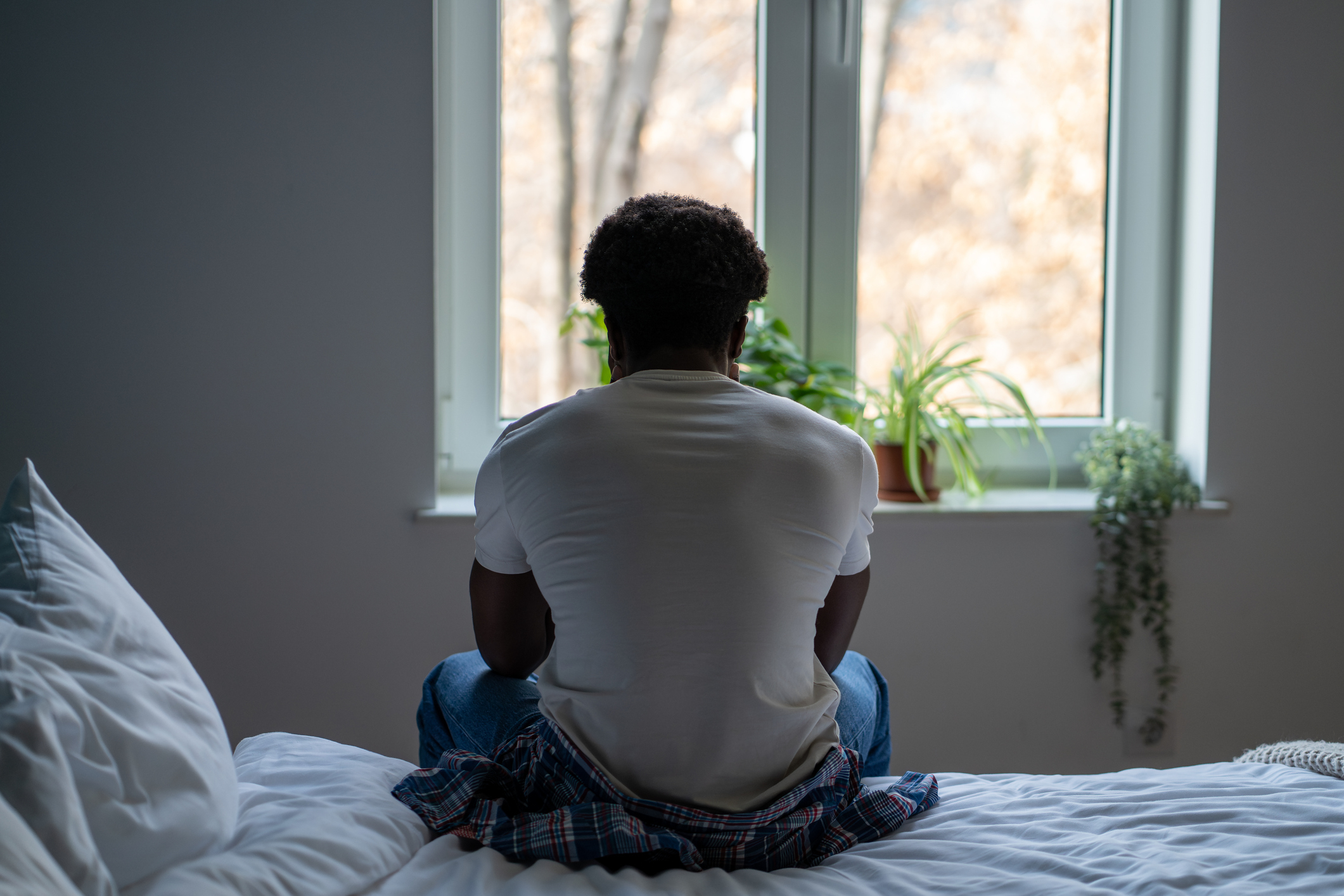 A person sits on the edge of a bed looking out of a window, appearing contemplative. Houseplants adorn the window sill