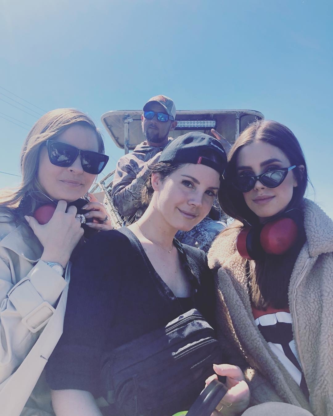 Lana Del Rey and two friends posing outdoors with Jeremy in a cap and sunglasses in the background