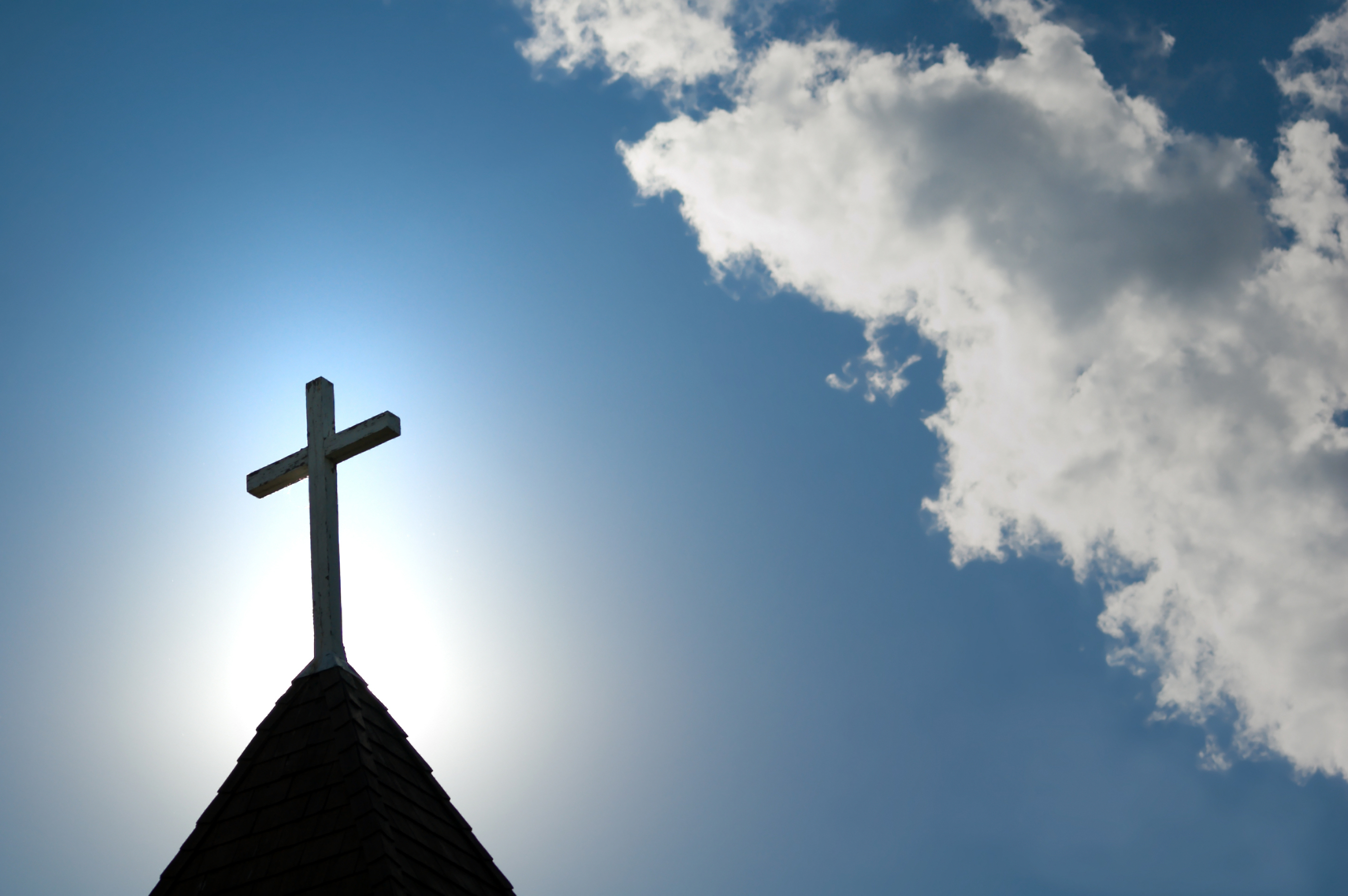 A church steeple topped with a cross is silhouetted against a bright sky with clouds