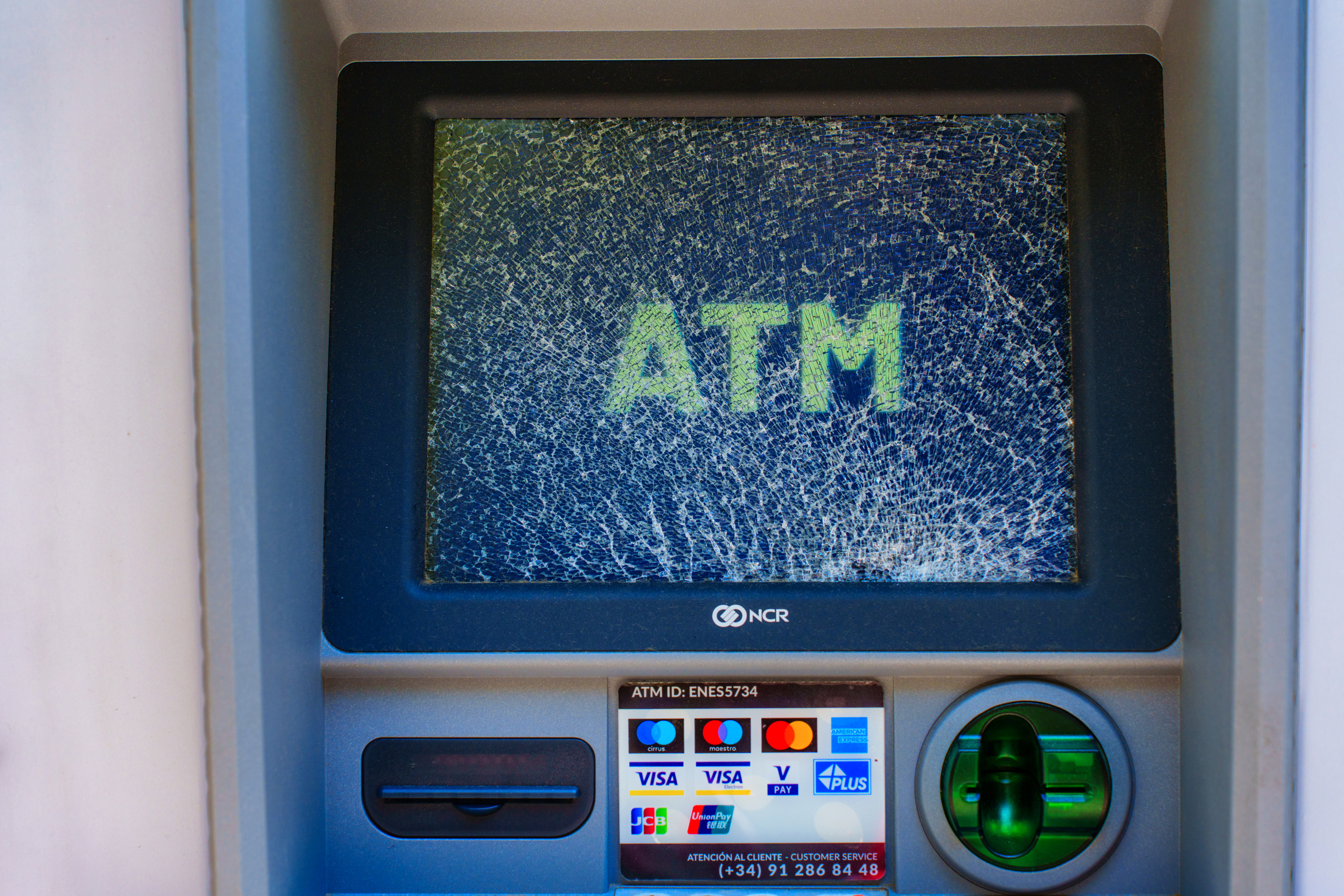 A damaged ATM screen displays the word "ATM" with a visible spiderweb crack. The card reader and keypad below remain intact