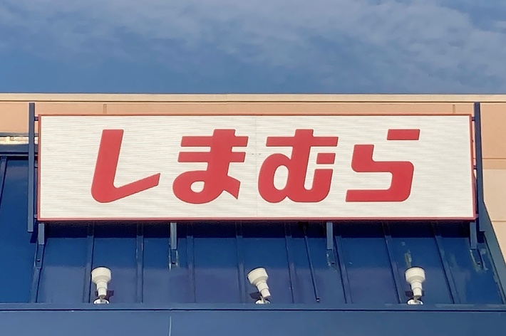The image shows a storefront with a sign in Japanese that reads “しまむら”. There is also a blue accessible parking sign in view