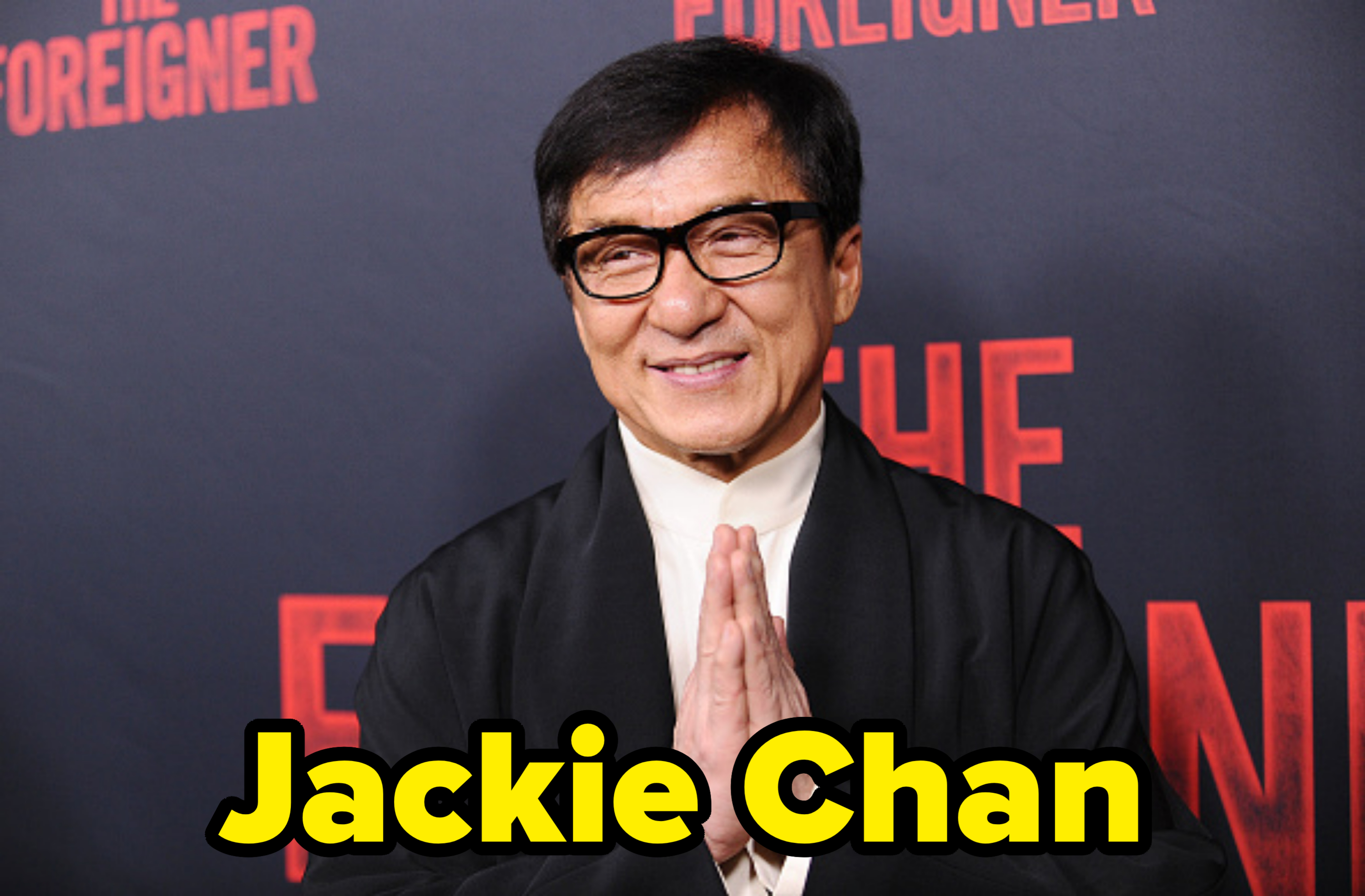 Jackie Chan on the red carpet for &quot;The Foreigner&quot; premiere, wearing a traditional outfit with a black robe over a white high-collared shirt, smiling with hands clasped
