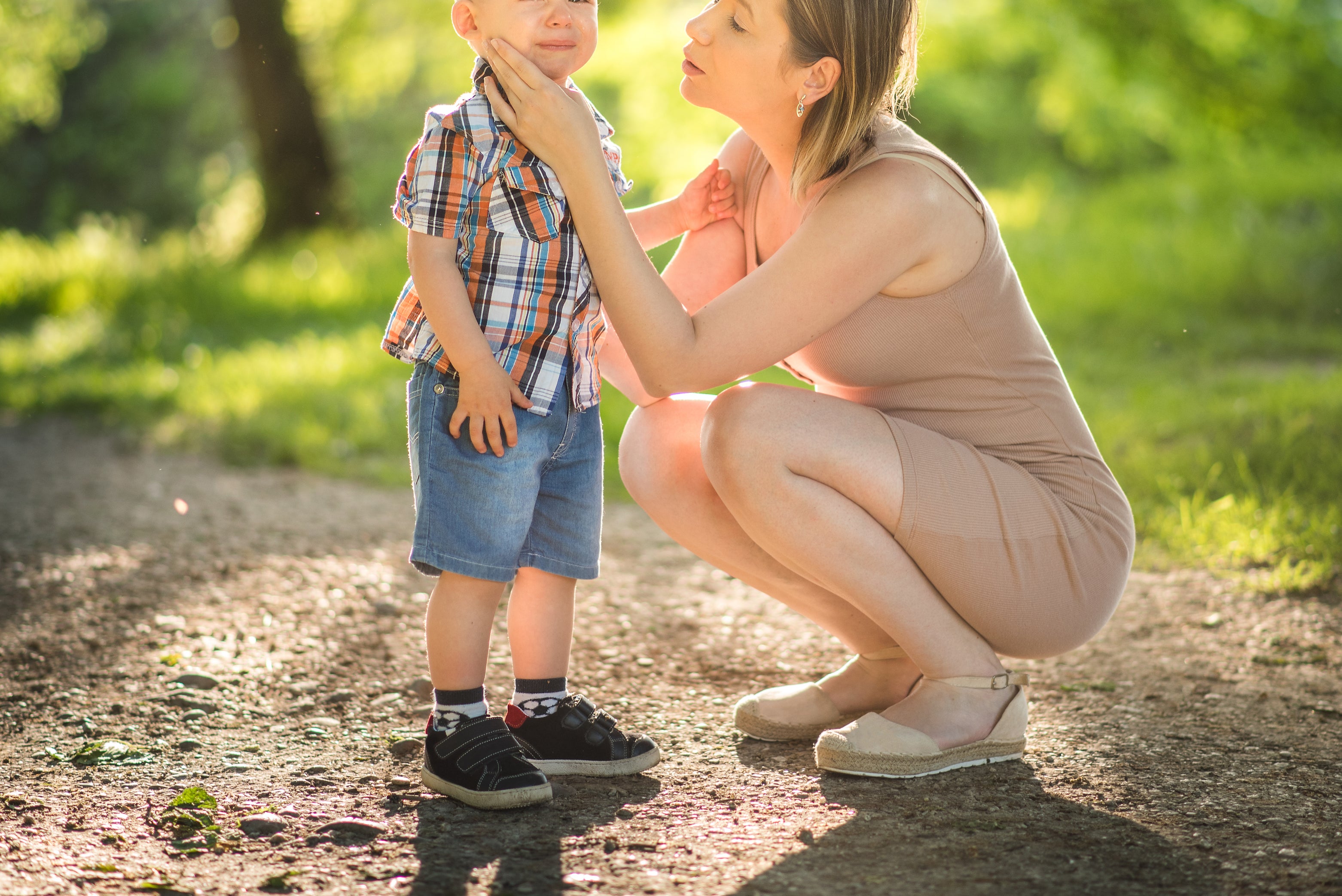 A woman lovingly cups the face of a smiling young boy in a sunlit outdoor setting. Trees and greenery fill the background.