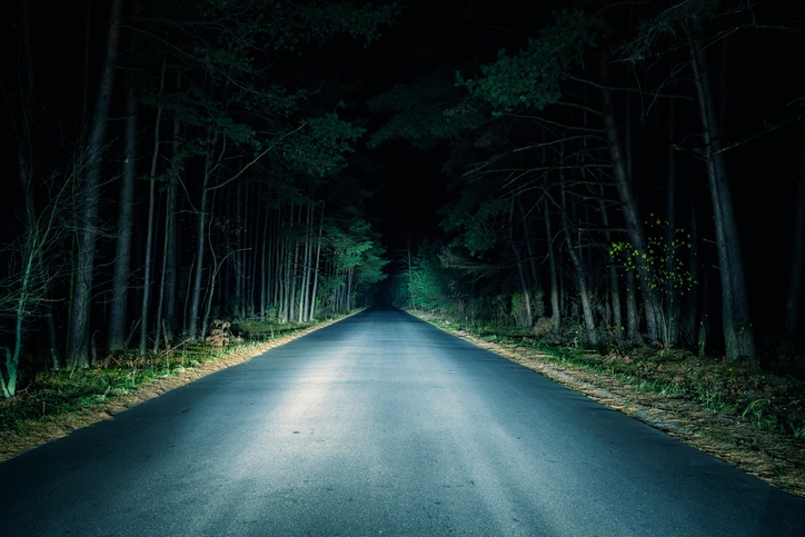 A deserted road cutting through a dark, dense forest at night, illuminated by headlights, creating an eerie and mysterious atmosphere