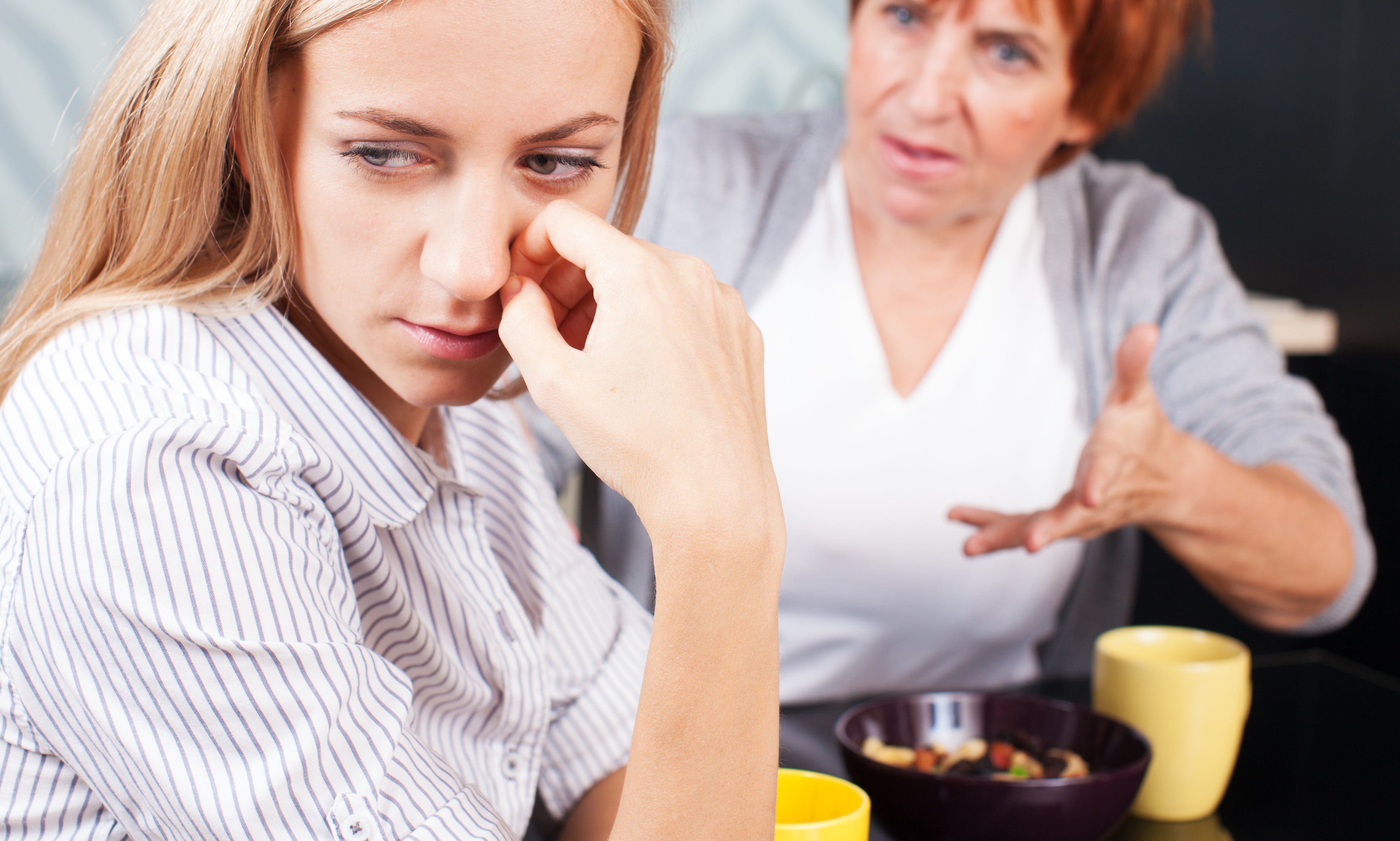 A woman with long blonde hair looking sad sits at a table while an older woman with short hair gestures emphatically towards her
