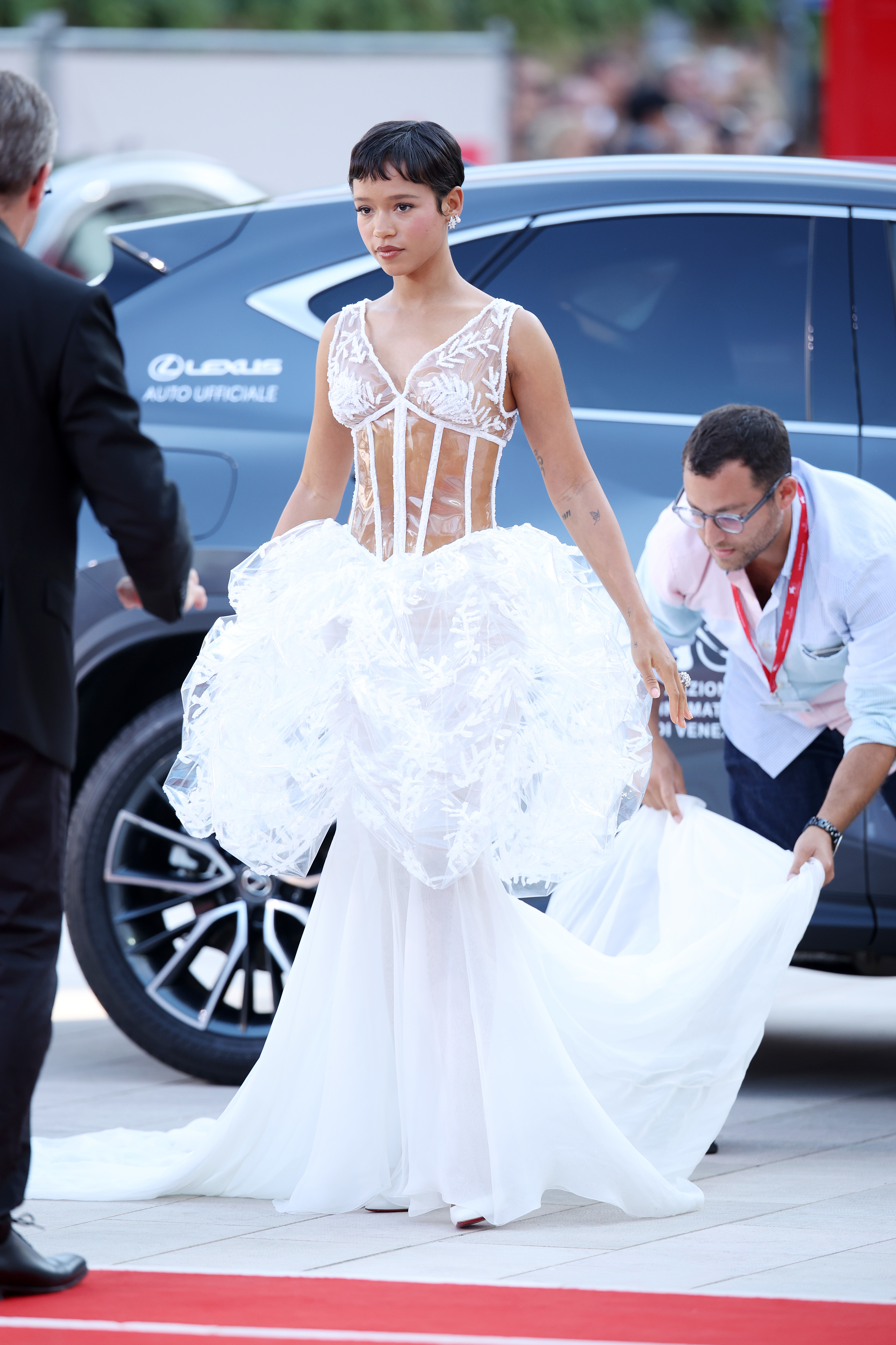 Taylor Russell wears a unique gown with a sheer bodice and voluminous skirt on the red carpet, accompanied by an assistant arranging her dress
