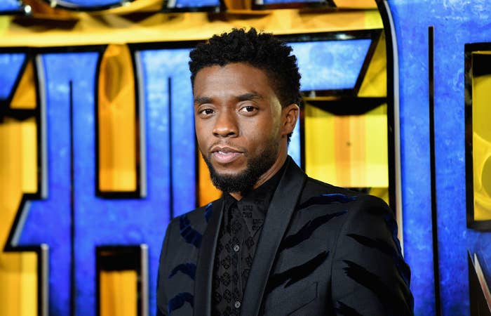 Chadwick Boseman stands in front of a movie premiere backdrop, wearing a patterned dark suit