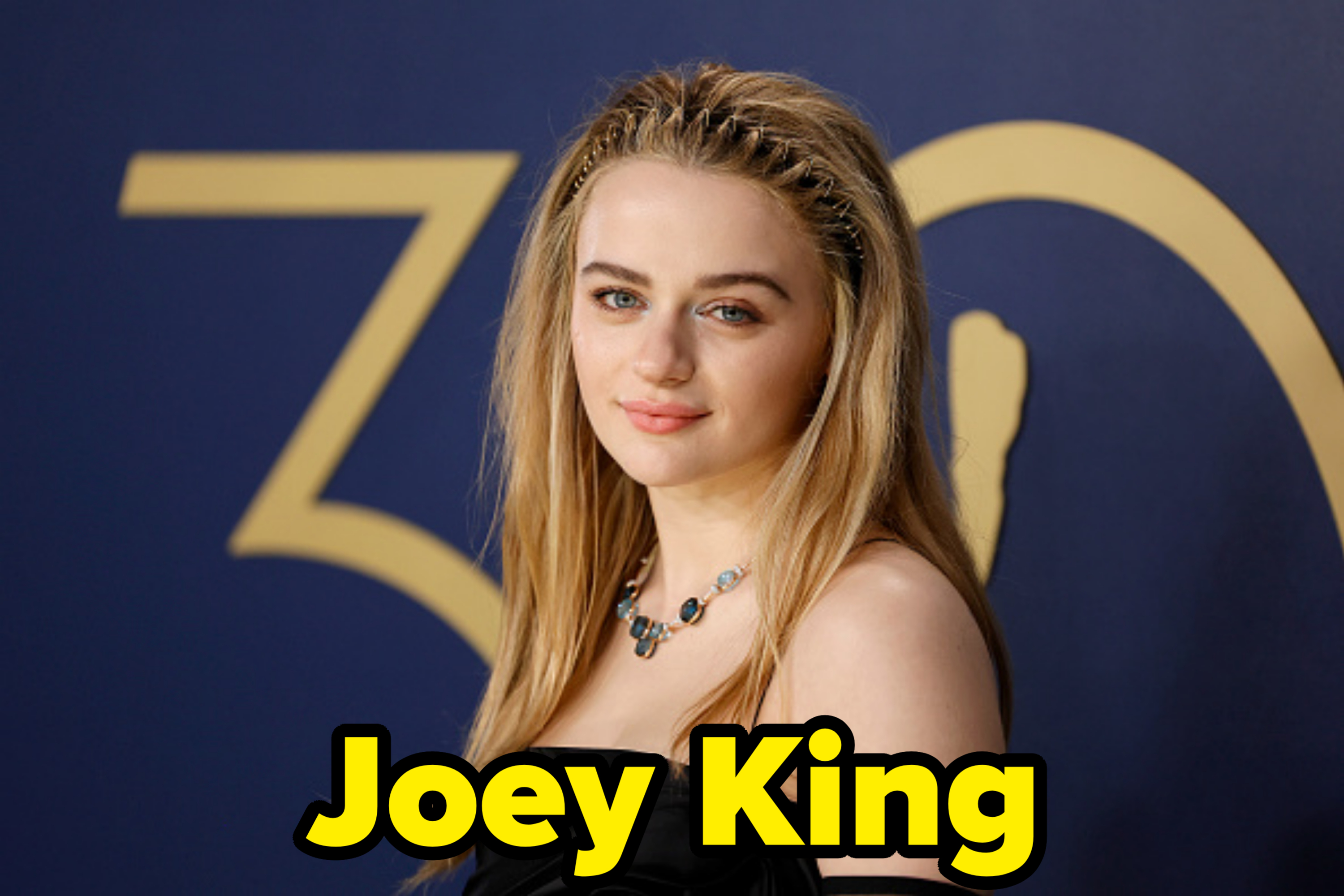 Joey King wearing a strapless black gown and gemstone necklace, posing against a backdrop with the number 30