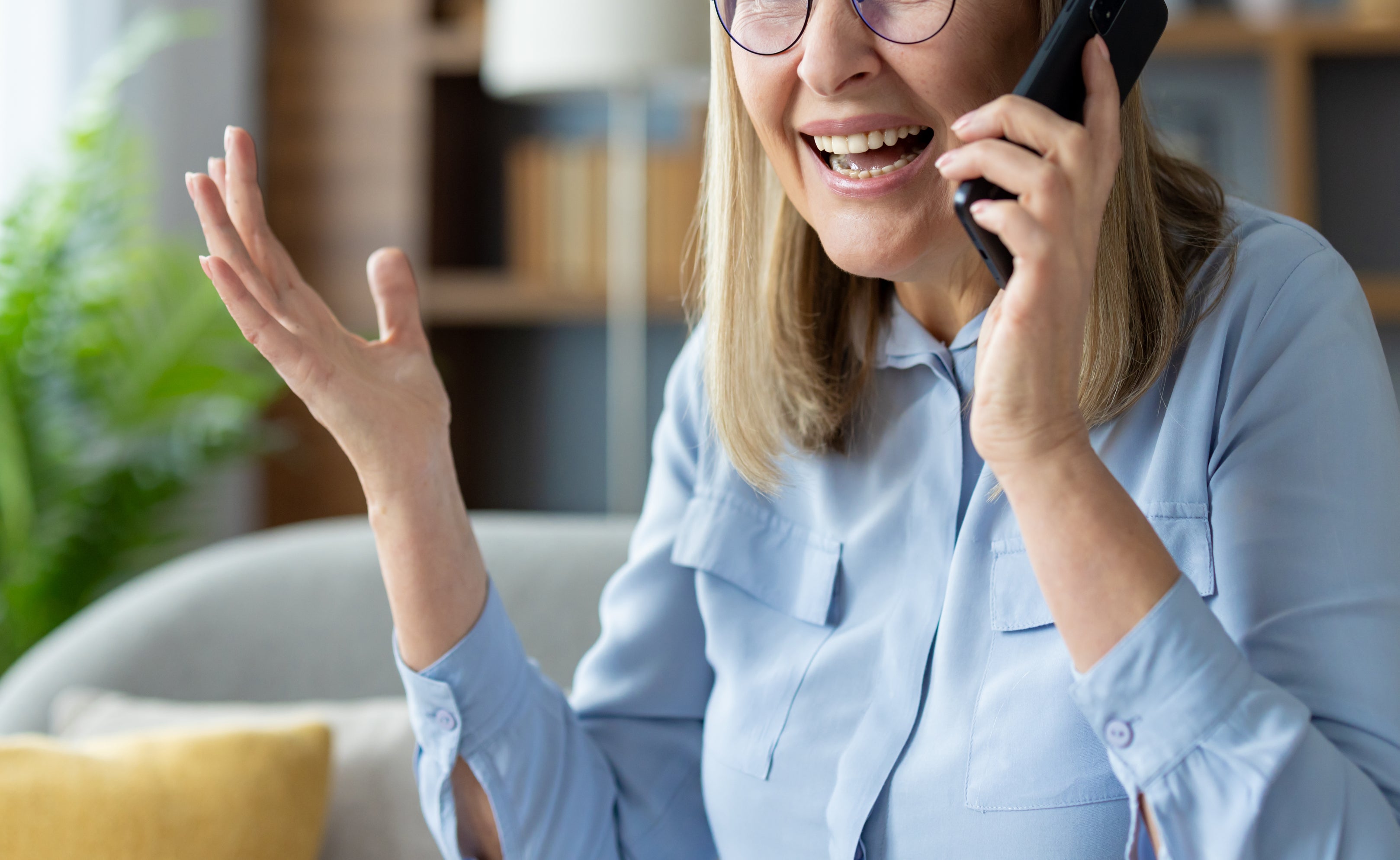 An elderly person with glasses and long hair talks expressively on a phone, appearing frustrated or animated, sitting in a living room