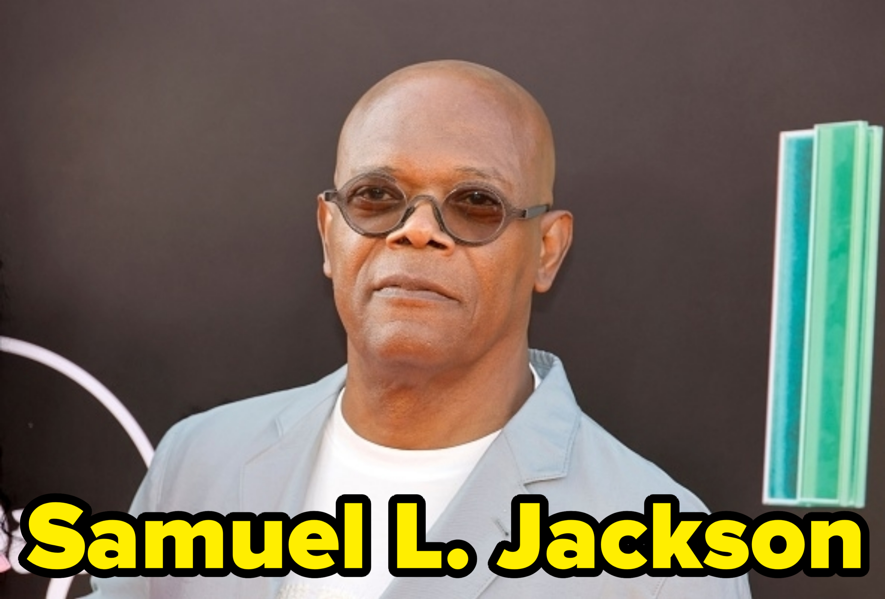 Samuel L. Jackson wears a light-colored suit and round sunglasses on the red carpet. He is posing in front of a dark backdrop