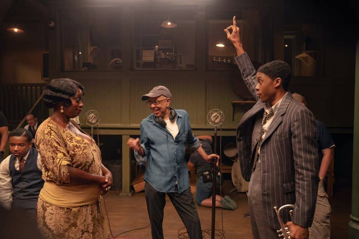 Viola Davis, Chadwick Boseman, and George C. Wolfe have a discussion on a film set. Chadwick holds a trumpet and raises his hand, while George gestures animatedly