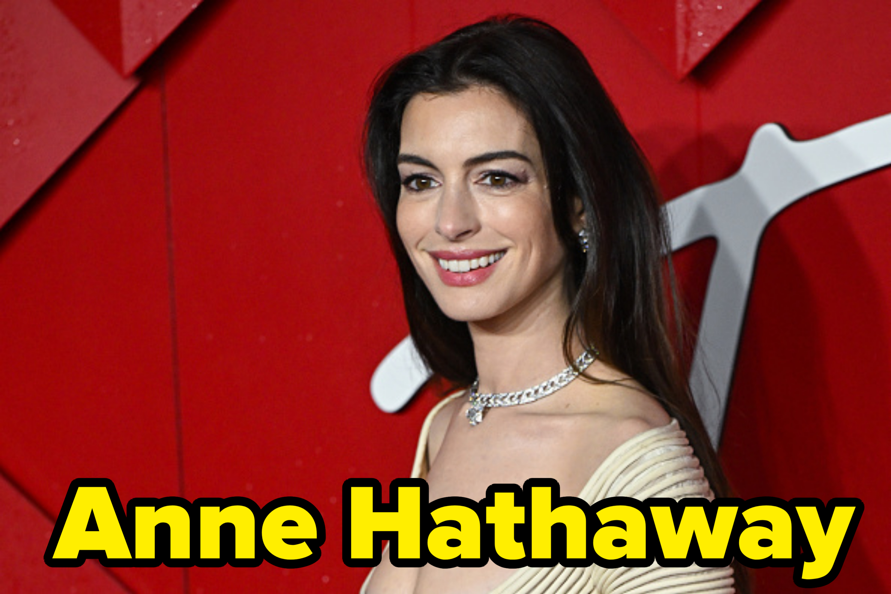 Anne Hathaway on the red carpet, wearing an elegant off-the-shoulder dress with a diamond necklace, smiling towards the camera