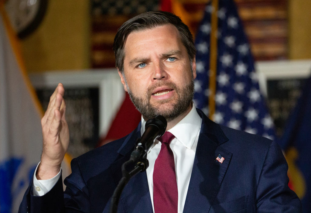 J.D. Vance speaking into a microphone, gesturing with his right hand. Multiple American flags are visible in the background