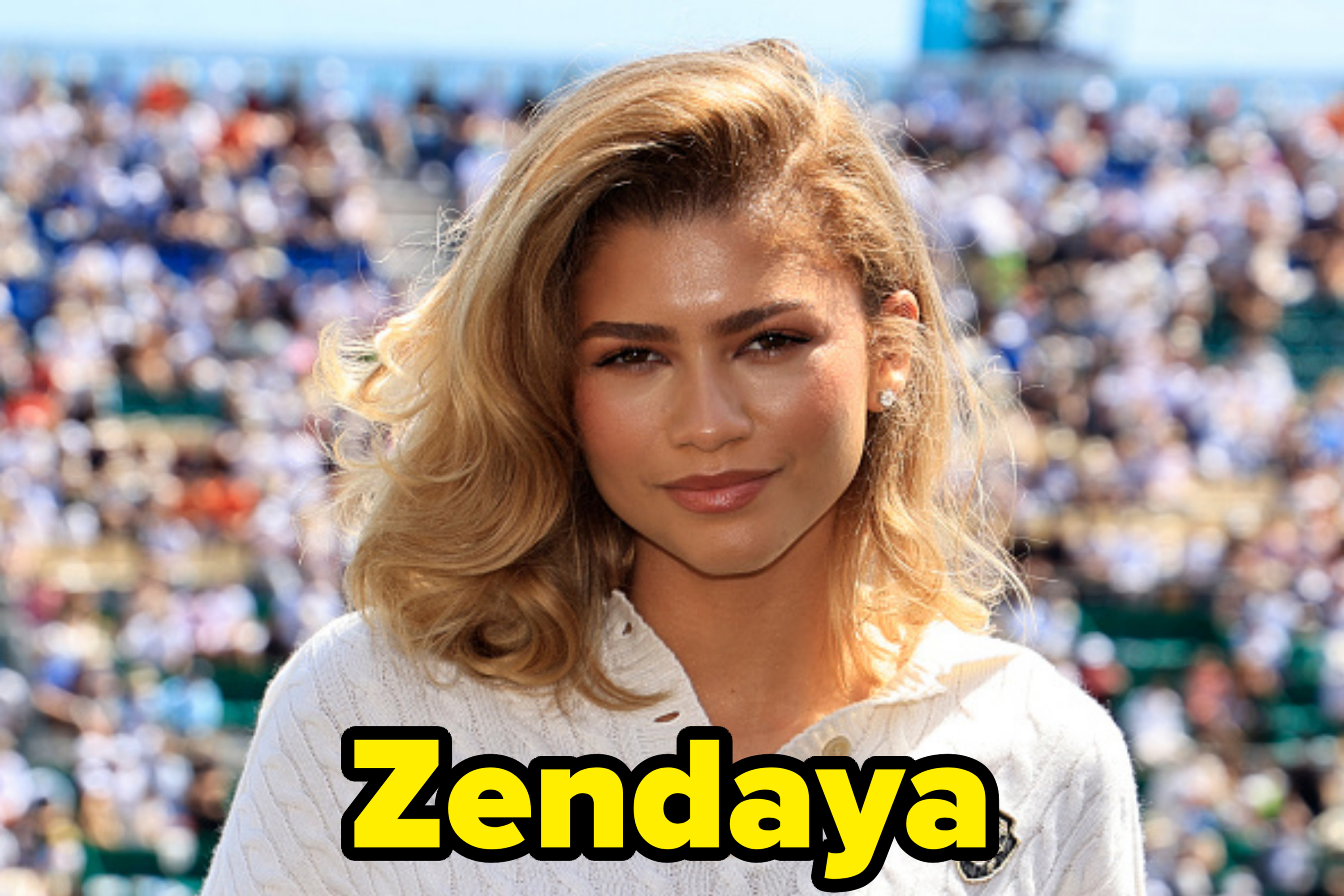 Zendaya at a sports event, wearing a white knit sweater with a detailed crest on it. The background shows a blurred crowd of spectators in the stands
