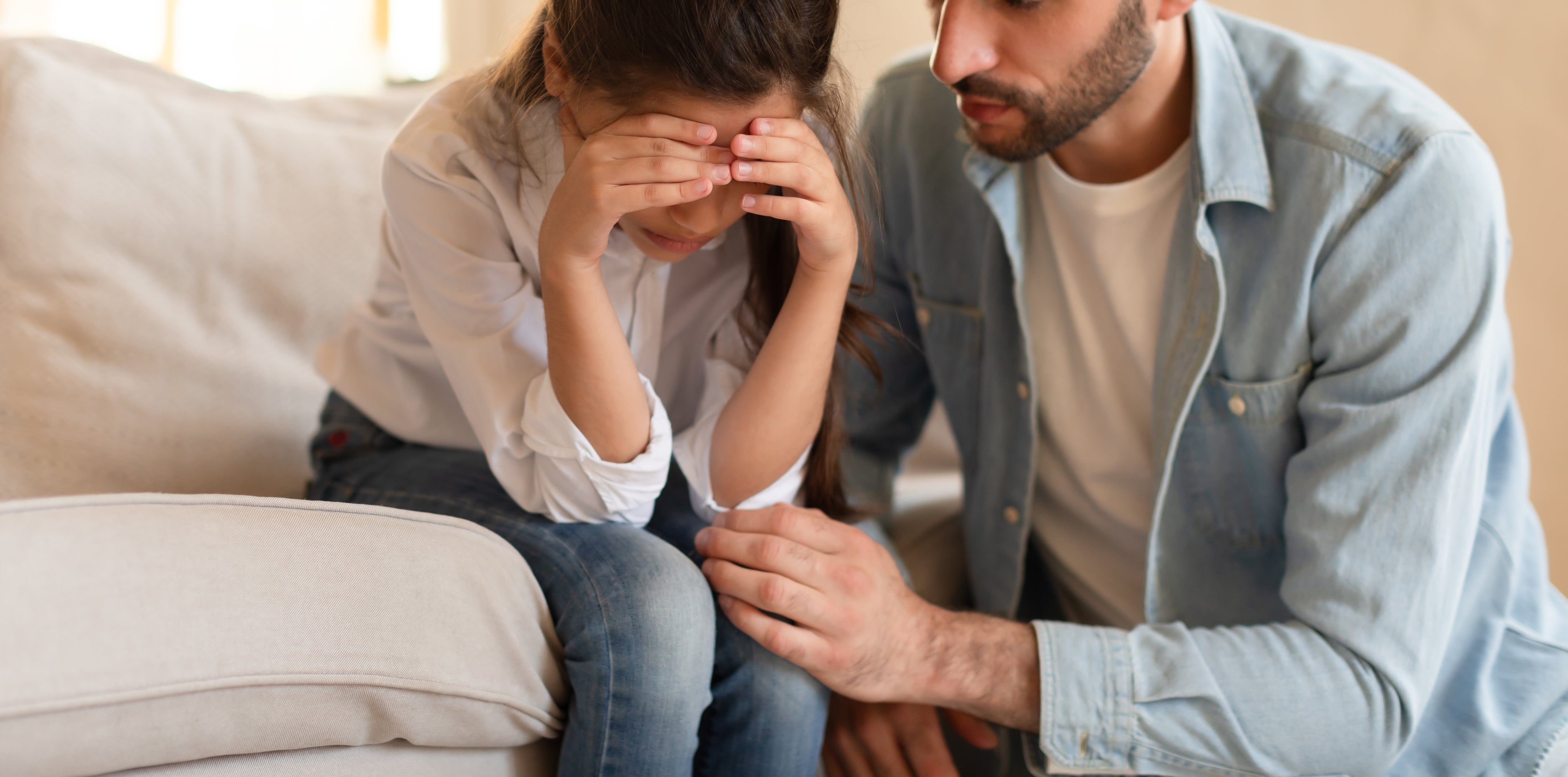 A man in casual clothing comforts a young girl who is seated on a couch with her hands covering her face. They both look concerned