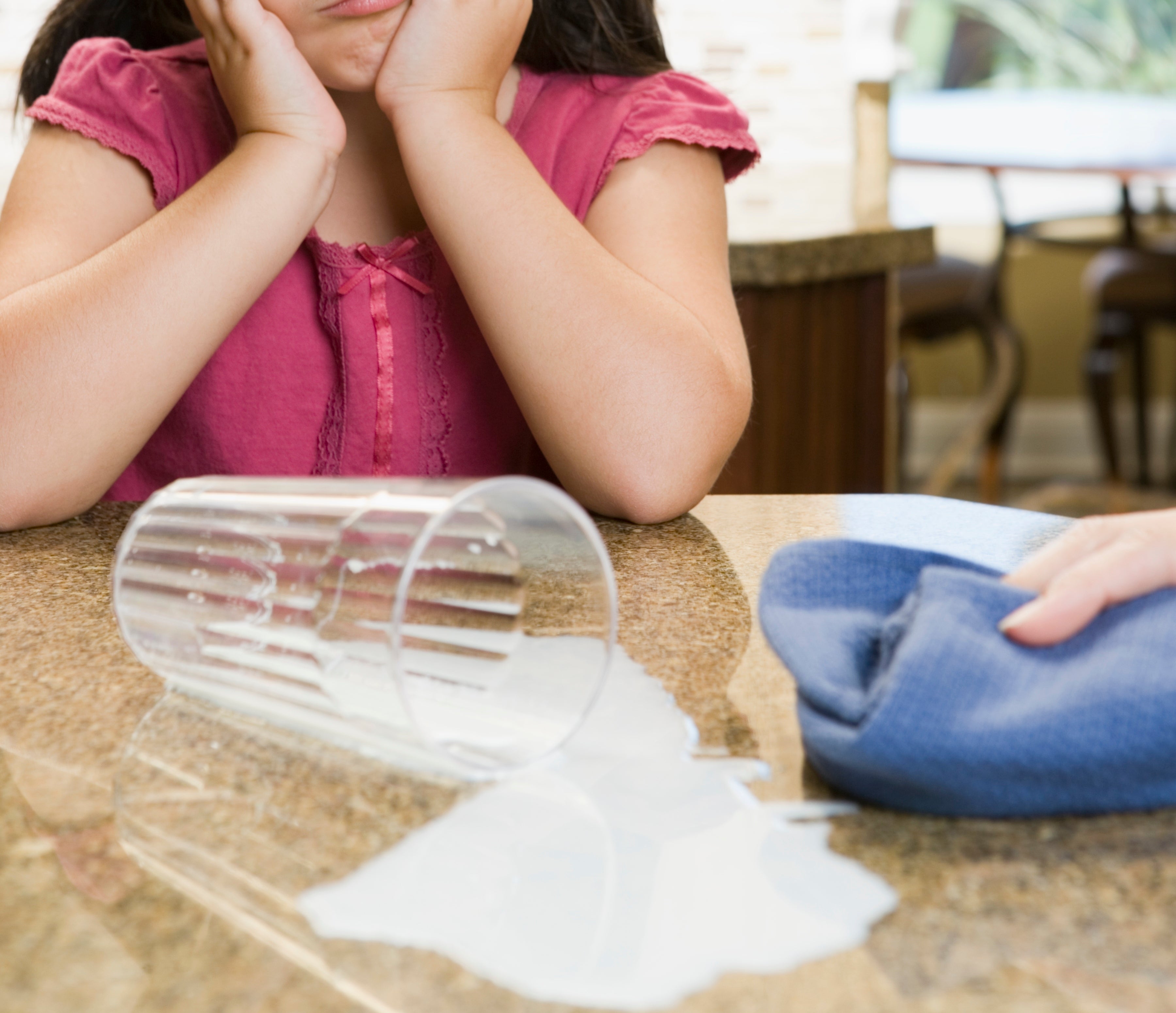 Young girl with a pensive expression, hands on her cheeks, looks at a spilled glass of milk on a kitchen counter, while an adult's hand holds a blue cloth