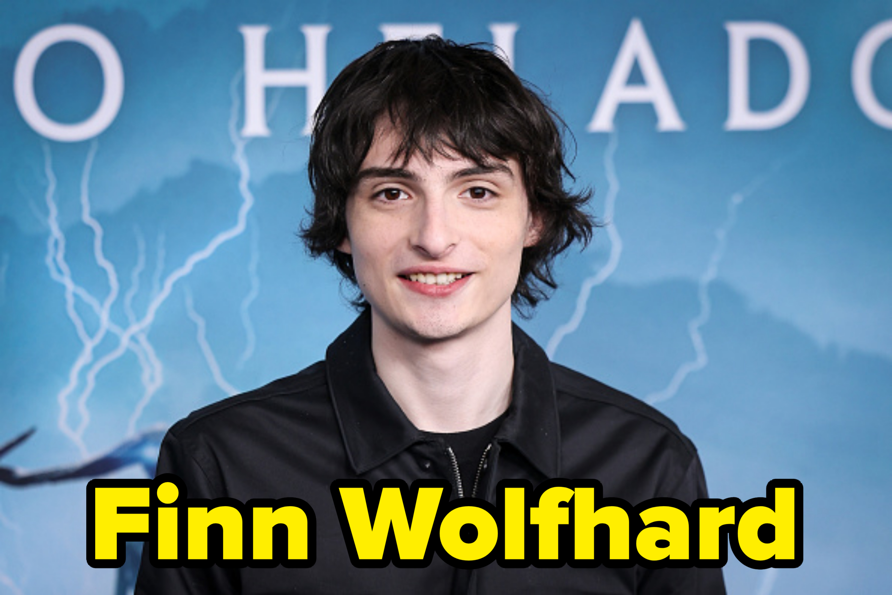 Finn Wolfhard, wearing a black jacket, stands in front of a blue background with lightning design at a public event
