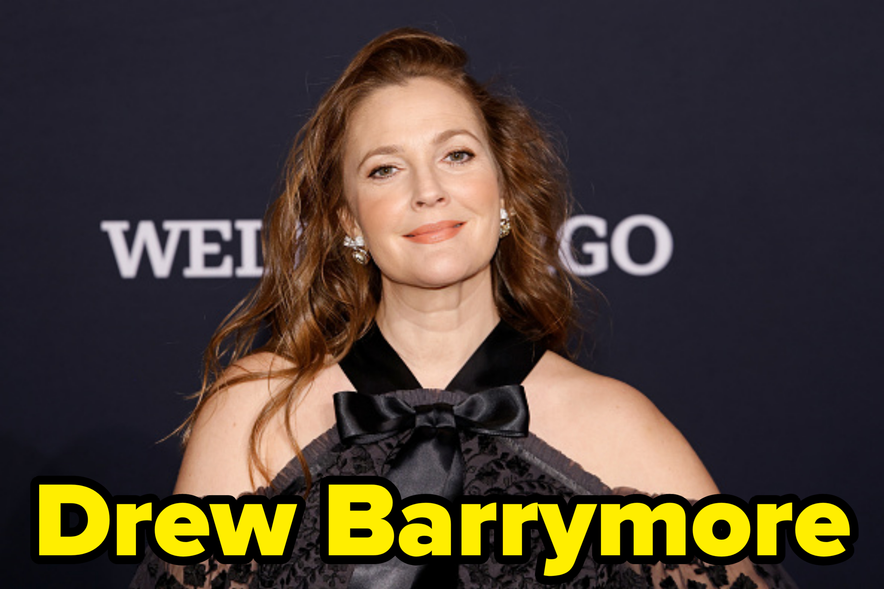 Drew Barrymore at an event, wearing an elegant off-shoulder black gown with lace details and a large bow at the neckline