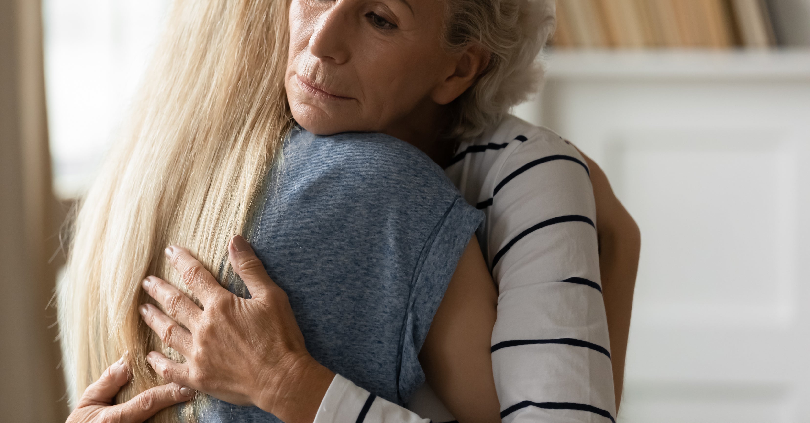 An older woman with curly hair embraces a younger woman with long blonde hair in a comforting hug, both appearing emotional