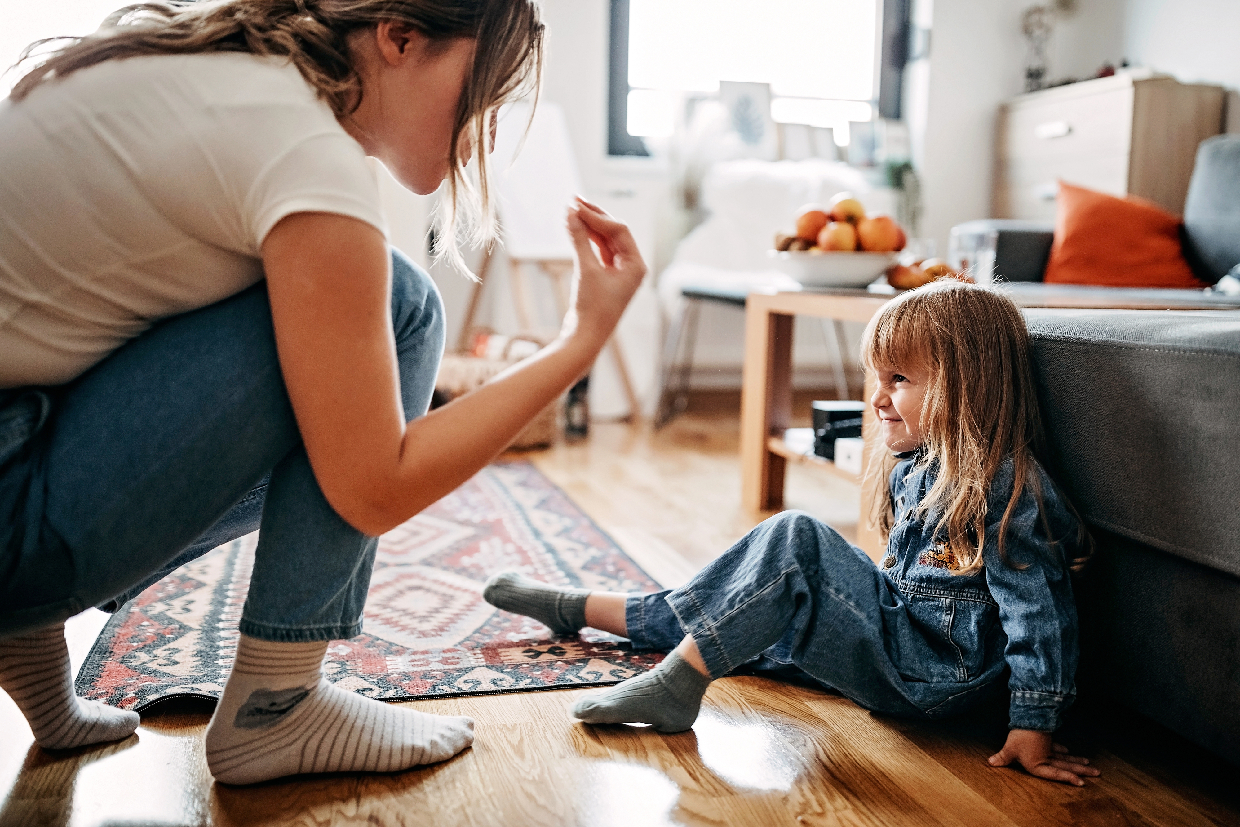 Woman kneeling and talking to a smiling young girl sitting on the floor in a cozy living room