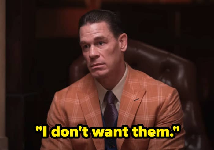 John Cena sits in a brown leather chair wearing a checkered suit and a tie with a concerned expression on his face