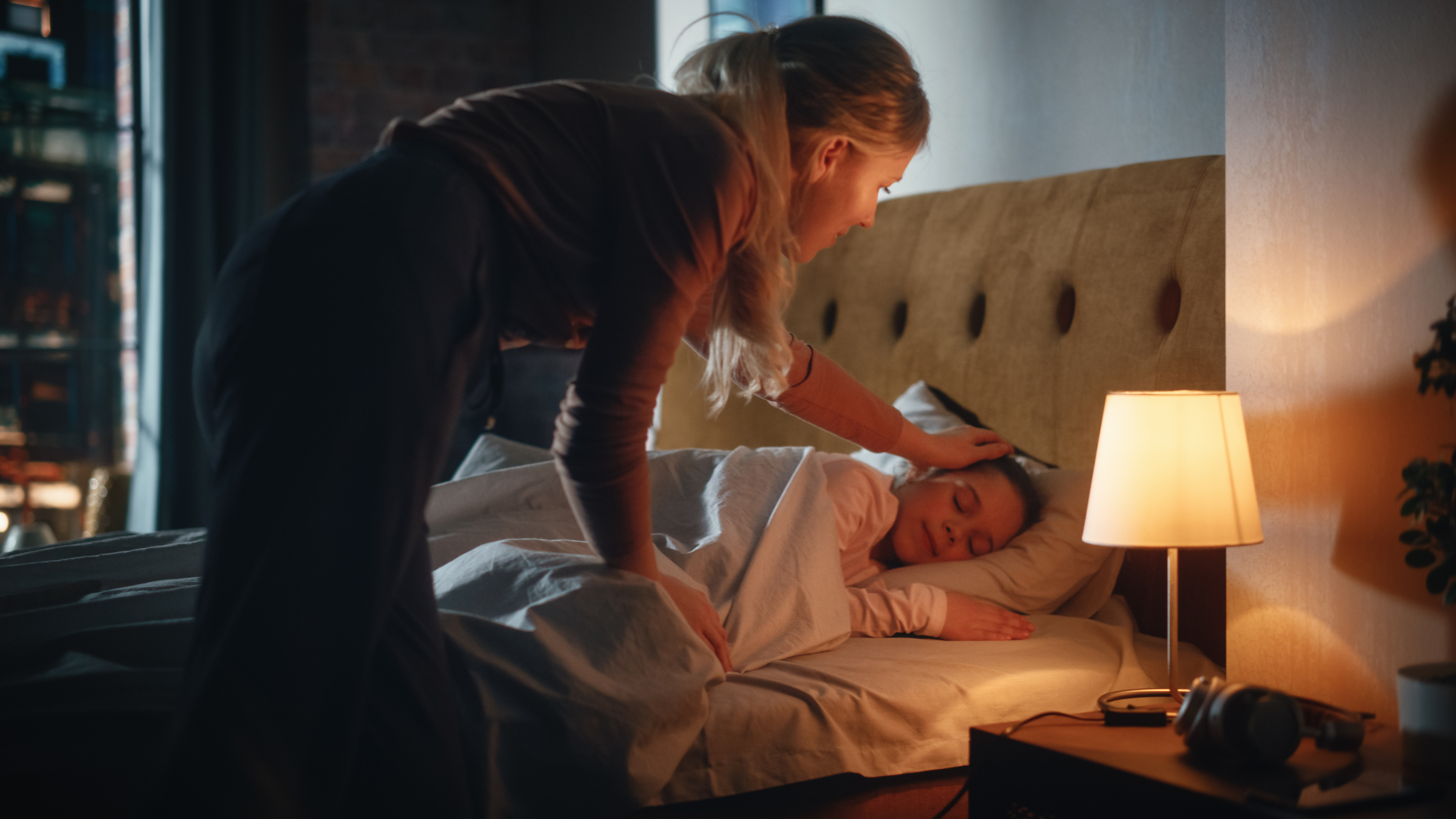 Woman tucks a sleeping child into bed, gently touching their forehead. A lamp is illuminated on the nightstand