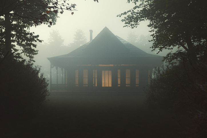 A small, illuminated house with a triangular roof is surrounded by dense fog and trees in a forest setting