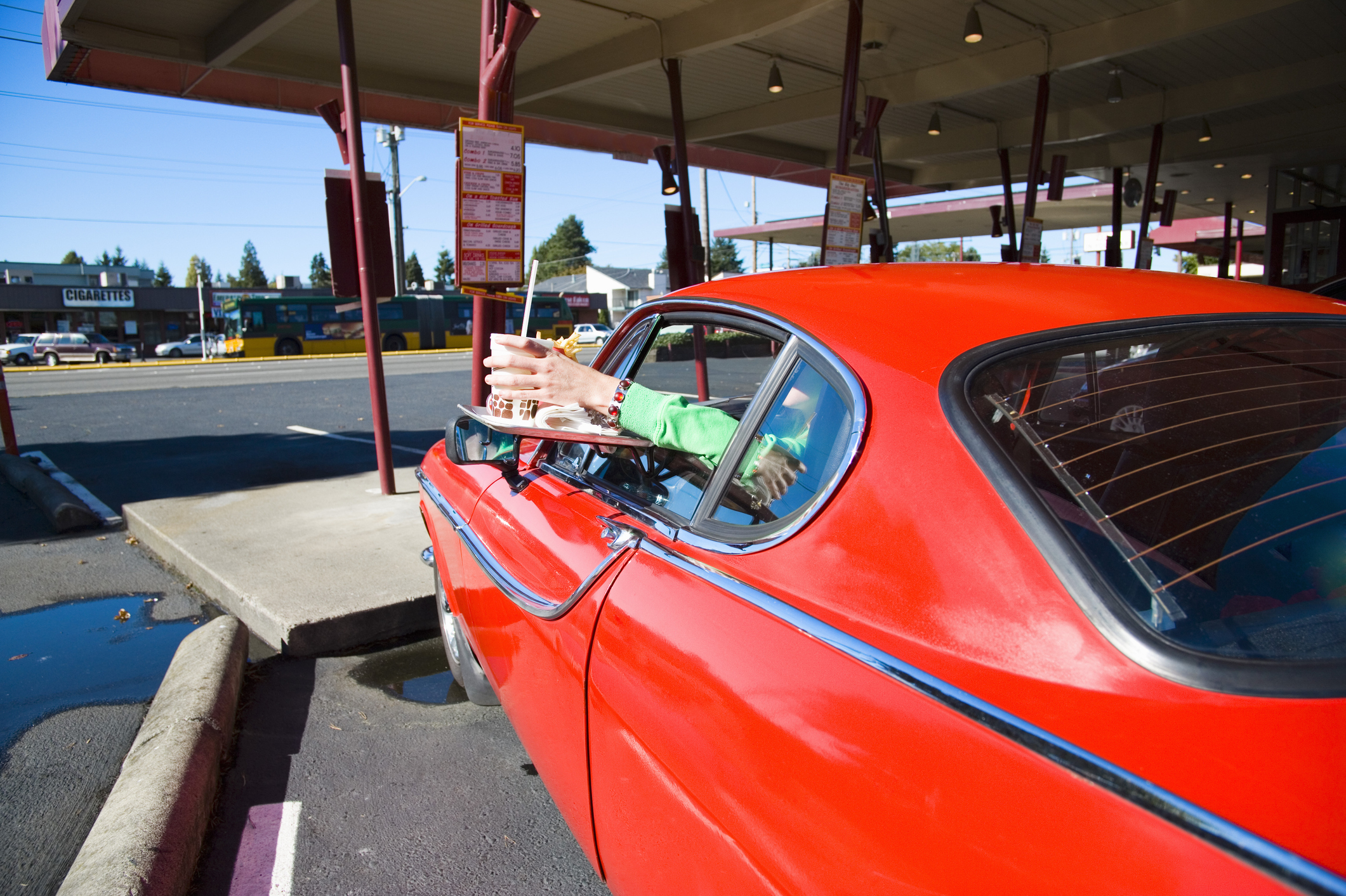 A classic car pulls up to a drive-in diner, with a person extending their arms out of the car window to receive service. The scene evokes nostalgic vibes